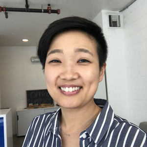 Angie Han is an Asian person with short black hair, buzzed on one side. Angie has a big smile and a blue and white stripped collared shirt, and is standing inside of what looks like a living room, taking a selfie. The image is a circle on this section of the website.