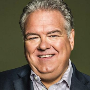 Jim O'Heir is a white man with gray and white hair. He has a big smile, large cheeks, and a stripped collared shirt with a dark gray jacket over it. The picture is a circle, and the background of the headshot is dark green.