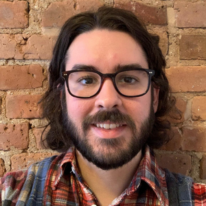 Liam Matthews is a white man with facial hair, and shoulder length wavy dark brown hair. He has on black glasses and long eyelashes. He is standing in front of a brick wall, and has on a collared plaid colorful shirt.