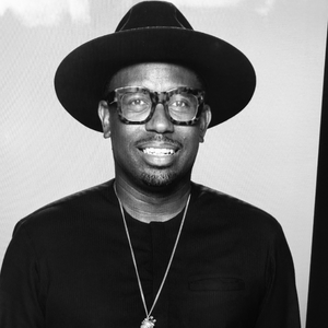 Mikael Moore is a Black man, and this is a black and white portrait. He has on a black hat and large rectangular tortoise glasses, a black long sleeve shirt, and a dog tag. He has some facial hair, and is smiling in the photo.