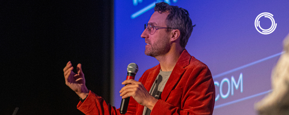 A white man with some facial hair and short brown hair is speaking with passion to an audience. He is holding a mic and standing in front of a screen projecting a purple background, holding a microphone, and wearing an orange velvet blazer and tan t-shirt.