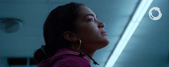 A Black woman with her hair back in a bun and gold hoops on looks off into the right of the camera. The camera is looking up, she is wearing a pink jacket, and the background is blue-gray.