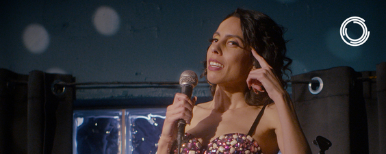 A thin brown woman with curly dark brown hair is standing right of center in the image. She is pointing to her head with one hand, and in the other holding a microphone. She is wearing a bejeweled top with black spaghetti straps, looking like she is performing. She is standing in front of a blue wall and a window, with black curtains open.