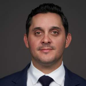 Travis Tammero is a brown man with light facial hair. He has gelled back dark hair and a black suit with a white collared shirt and black tie on. He's posing for a professional headshot in front of a gray background.
